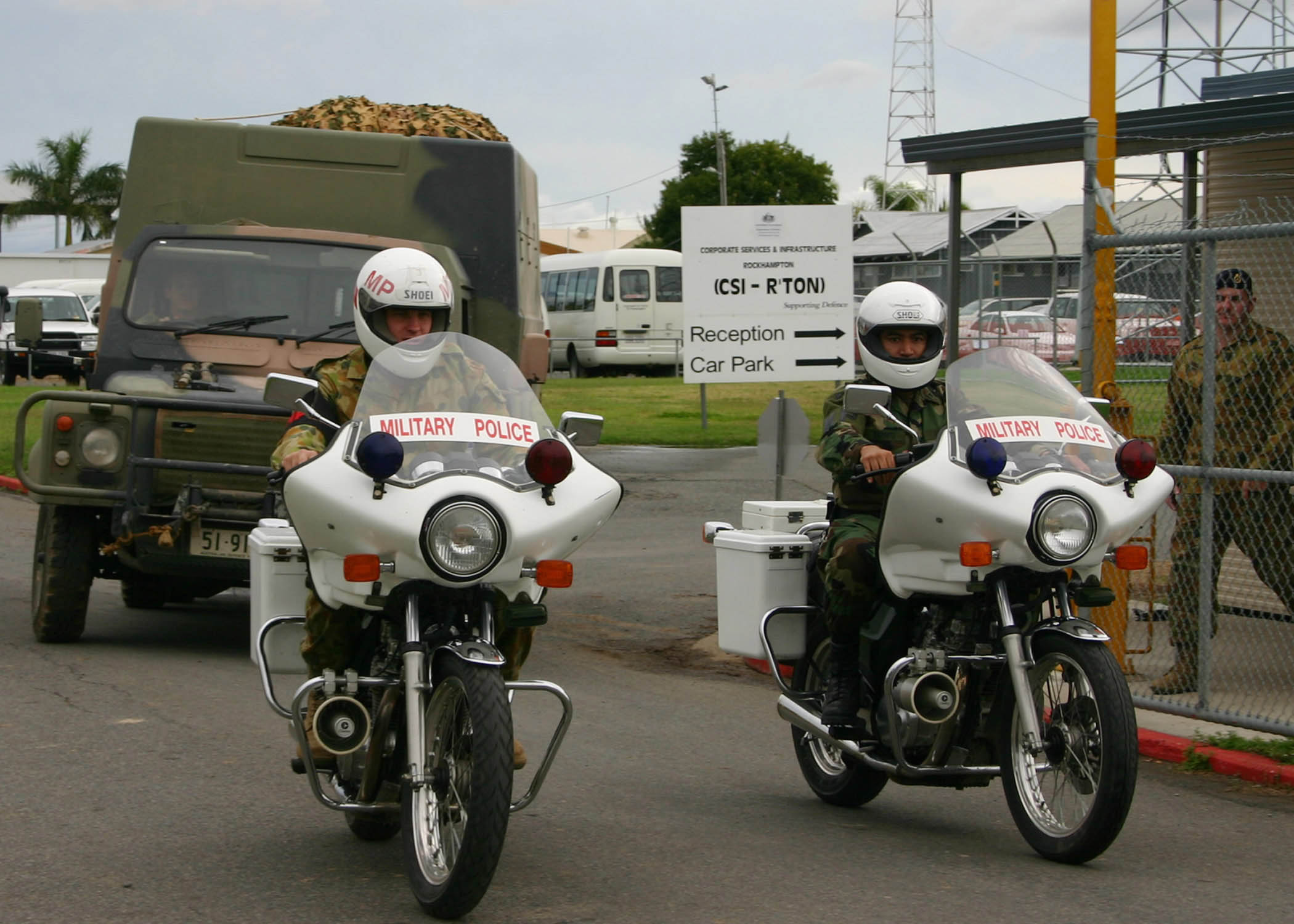 A Perentie 110 4×4 carrying a tall moulded hard rear body instead of the fleet's usual canvas tilt, behind Australian Army military police motorcycles at Camp Rocky, Queensland, June 2005. The source caption does not identify which enclosed-body variant it is, and no freely licensed photograph of a Perentie Survey panel van, Carryall or Command Post has been found.