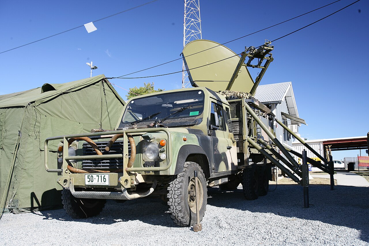 A Perentie 6×6 (ARN 50-716) in Auscam with its cargo tray uncovered, carrying an ADF Satellite Terminal Assembly during Exercise Talisman Sabre at Camp Rocky, Queensland, July 2009. The tilt and hoops are off — this is the same open tray the cargo variant carries in the general service role, shown here under a specialist load.